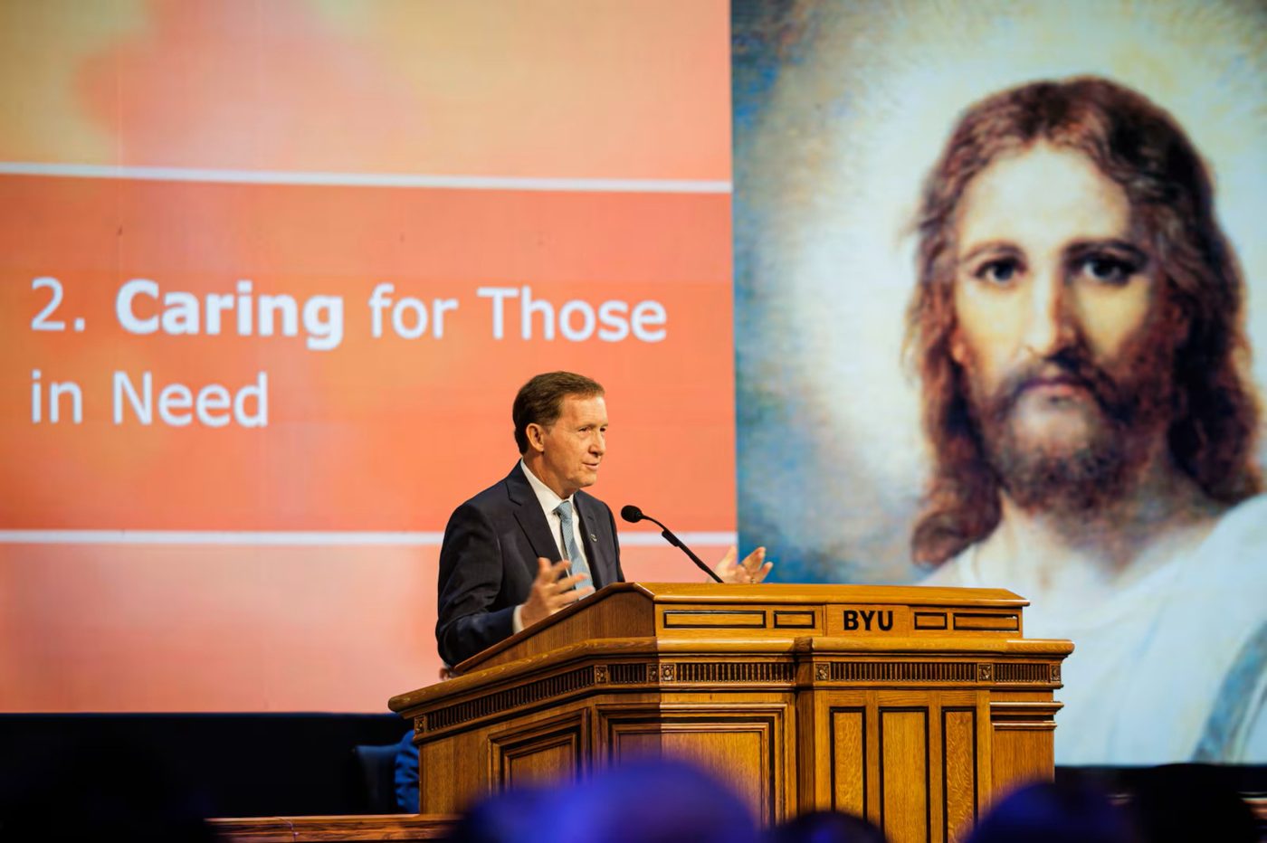 Bishop L. Todd Budge speaking at a BYU devotional pulpit beside a painting of Jesus Christ
